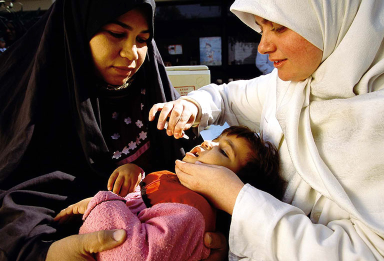 Two women in headscarves give medicine to an infant. One holds the baby while the other administers oral medication. © UNICEF/UNI21671/Pirozzi