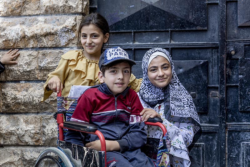 Mahmoud, 8, after attending summer remedial classes, supported by UNICEF, at a school, in Soukari neighborhood, Aleppo city, Syria, on 10 June 2023. His two sisters stand behind him with their hands on the wheelchair he sits in.