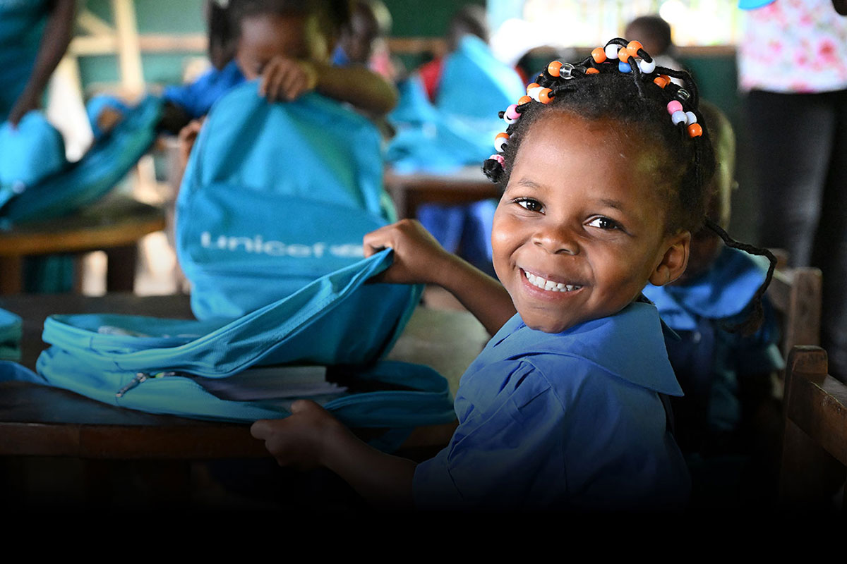 a young girl sits smiling at her desk, in a classroom full of children wearing blue UNICEF shirts with blue UNICEF backpacks. She sits reaching into her backpack.