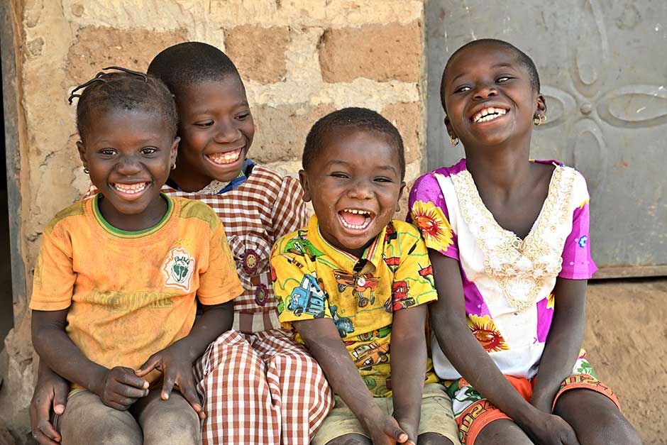 Four children sit smiling and laughing huddled together shoulder-to-shoulder on a brick building's stoop 