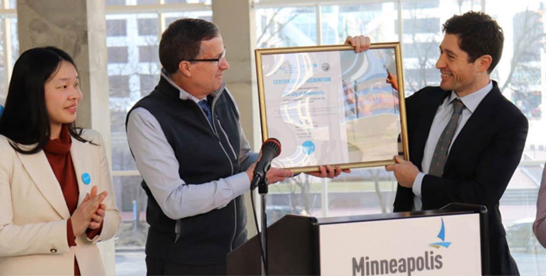 A UNICEF executive and a city official stand at a podium and jointly hold up a framed certificate celebrating Minneapolis’s designation as a child-friendly city.