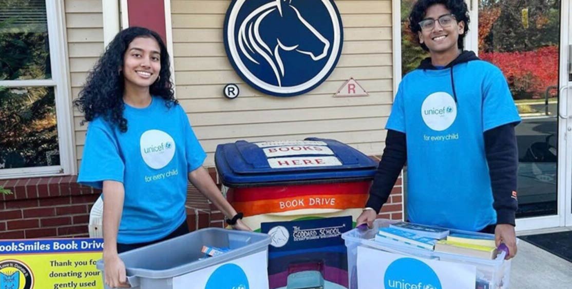 Two UNICEF club members hold clear plastic bins full of books and stand in front of a larger book bin that with a sign that reads 'Book Drive'.