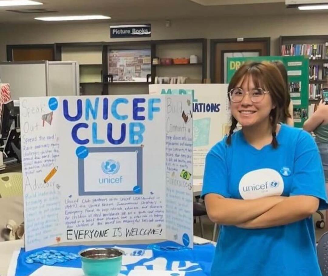 Two high school students cross their arms and smile next to their tri-fold poster display recruiting people to join their school’s UNICEF Club.