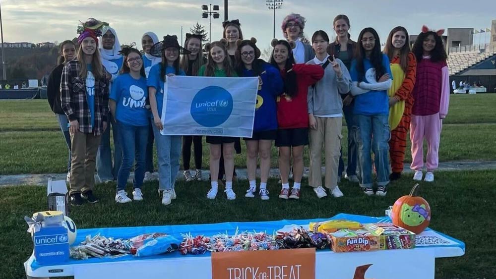 On an outdoor athletic field, a group of college students holds a UNICEF flag while standing behind a Trick-or-Treat for UNICEF table covered in candy.