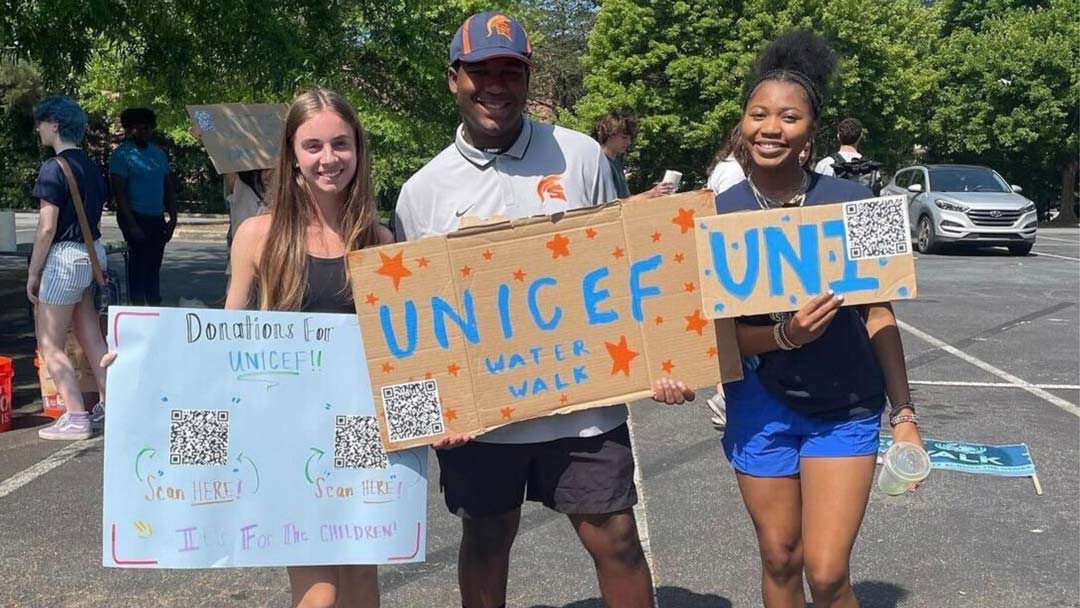 Three UNICEF club members stand in a parking lot in shorts and t-shirts and hold cardboard signs that announce their “water walk” and ask for donations for UNICEF through QR codes affixed to the sign.