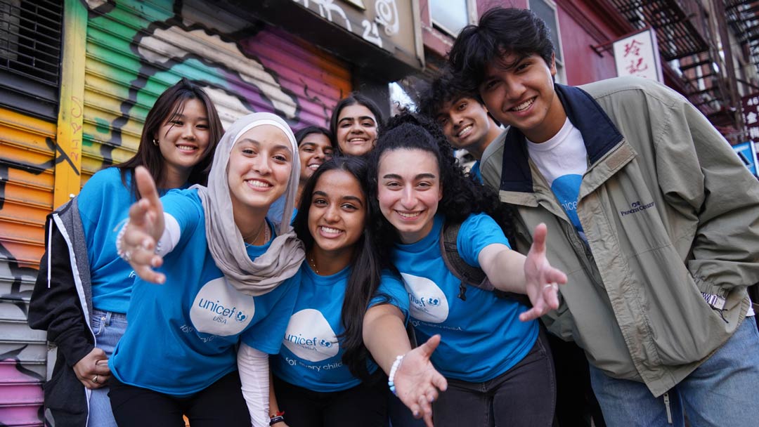 A group of smiling UNICEF club members, wearing UNICEF USA t-shirts, stand on a city street and reach their arms out to signify welcoming others.