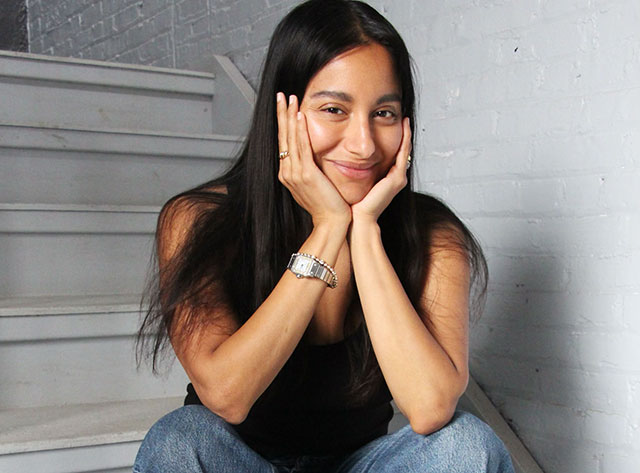 AMRIT sitting on stairs, resting her chin in her hands, eblows resting on her knees, and smiling.