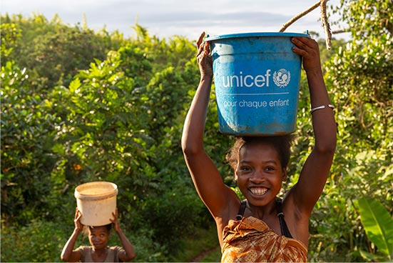 Two children carry buckets on their head as they walk outside in front of trees. The shorter child has a toy bucket on her head, and the taller child has a blue UNICEF bucket.