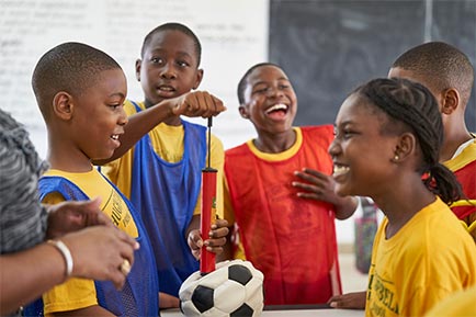 A group of children stand laughing in a classroom, one of them holding a football between them all.