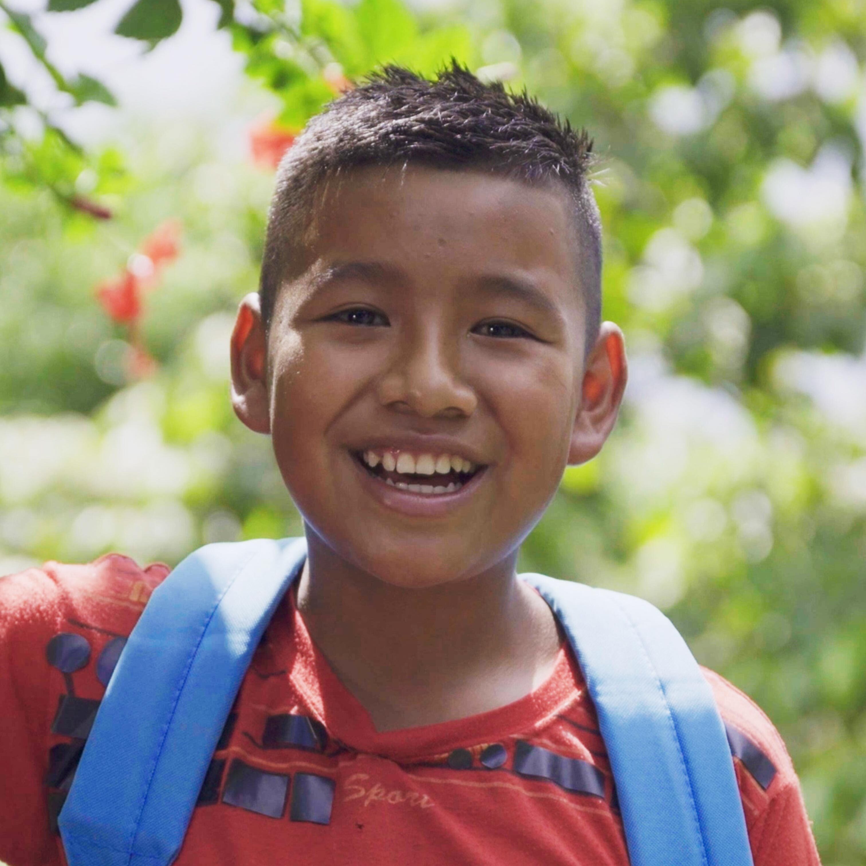 A portrait of Ludvin, a short-brown haired Guatemalan child, wearing an orange shirt and a blue UNICEF backpack, standing in front of trees outside and smiling.