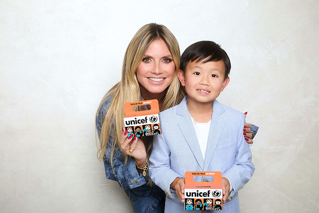 Heidi Klum leans down to pose with a child who's wearing a blue blazer. They both hold yellow UNICEF Trick-or-Treat boxes.