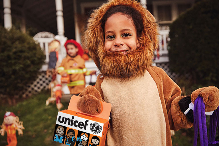 a laughing child wearing a lion costume holds up the Trick-or-Treat for UNICEF box.