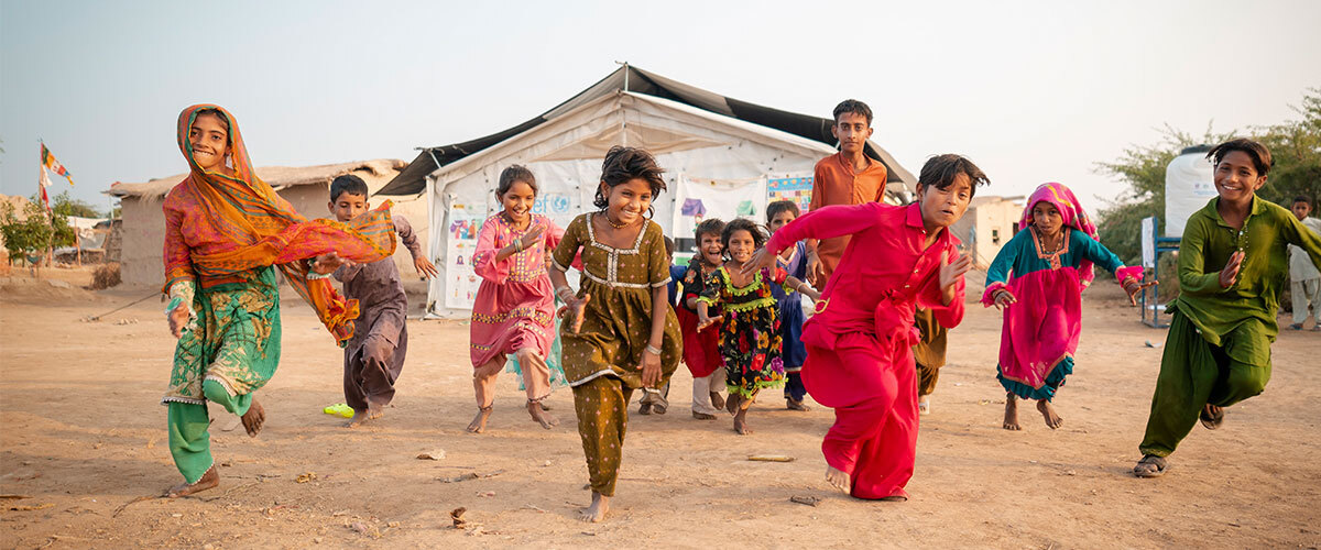 Children in ceremonial garb dance smiling in front of a UNICEF tent