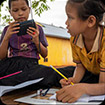 Two young students sit outside their school The boy looks at contents on his phone, and the girl is writing in a workbook.