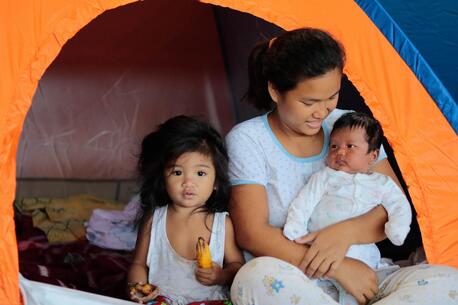 Woman with baby and small child cuddle in front of a tent in the Philippines following a severe tropical storm and displacement from their hoome