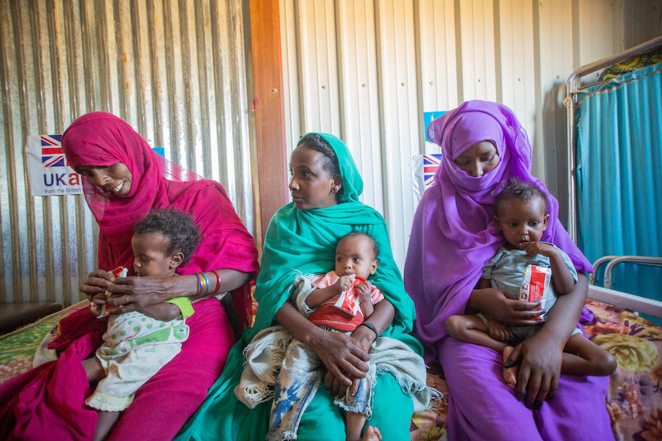 Three mothers sit side by side with their young children helping them eat Ready-to-Use-Therapeutic Food as part of their treatment for severe acute malnutrition at the Al-Arab health facility in Sudan.