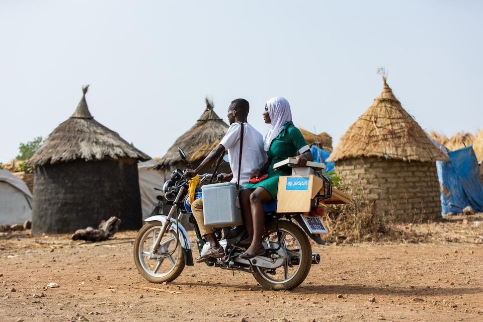 A UNICEF Immunization Officer and Community Health Nurse ride a motorcycle to carry vaccines in a cold box so they can provide immunization services to refugees at a camp in Sapelliga, Ghana.