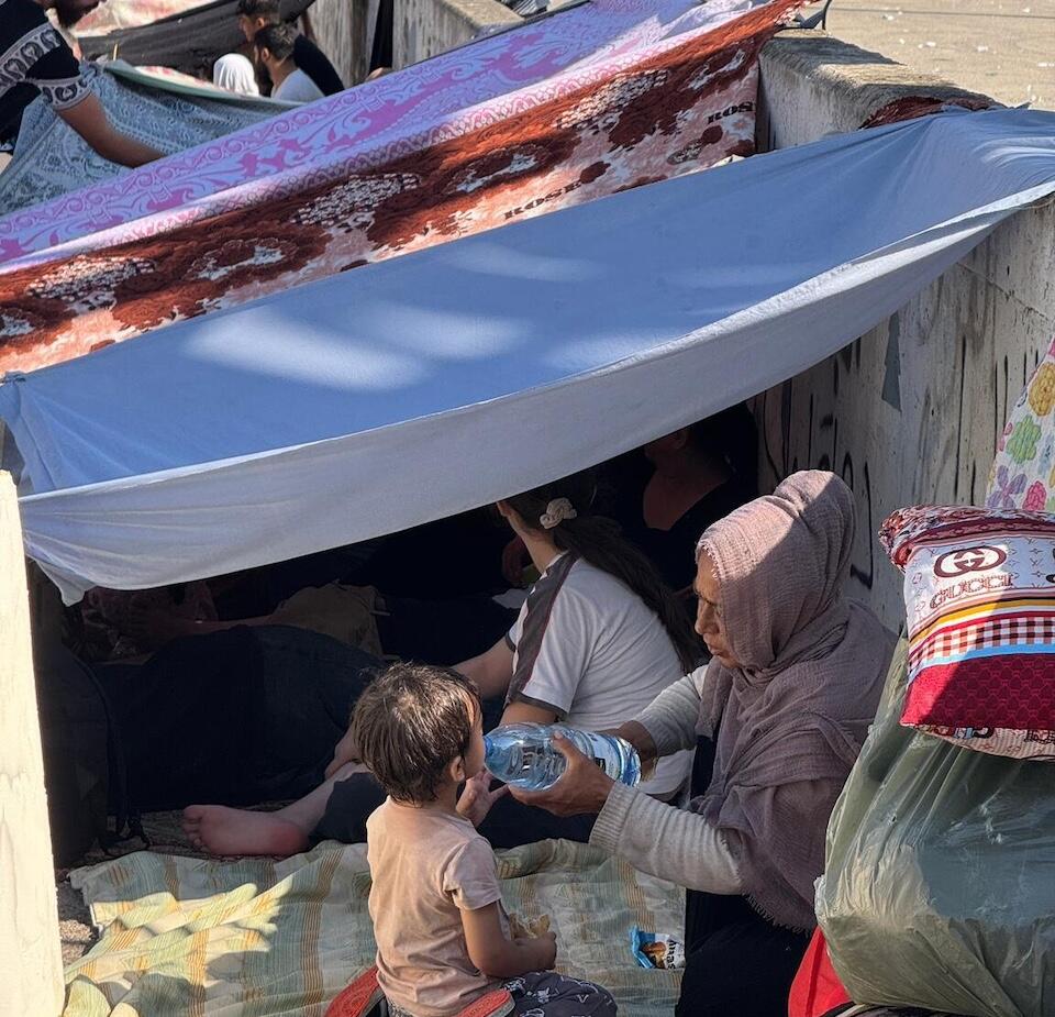 In Lebanon, next to a thin blanket serving as shelter for those displaced by conflict, a woman holds a bottle of water as a young child drinks from it. 