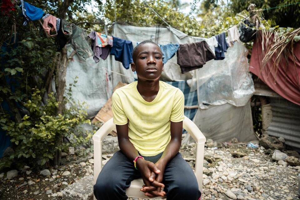 Makenley, 13, sits in a plastic chair in the corner of an IDP camp in Léogâne, southwestern Haiti, where he lives with his grandmother after losing both parents.