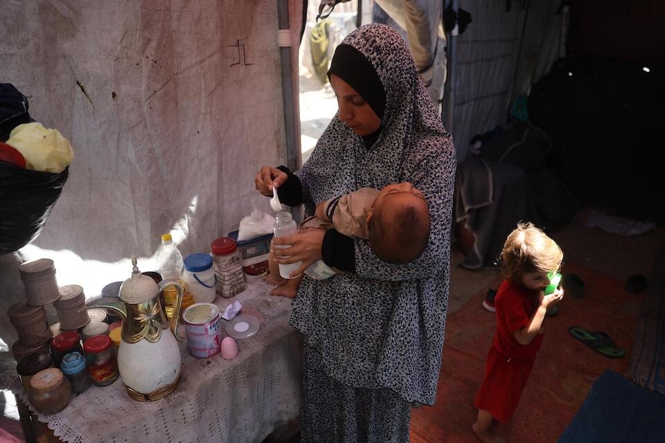 Madeline, a mother from Gaza, holds her child in her arms while preparing the milk for her baby. She and her family are beneficiaries of UNICEF’s cash transfer program.